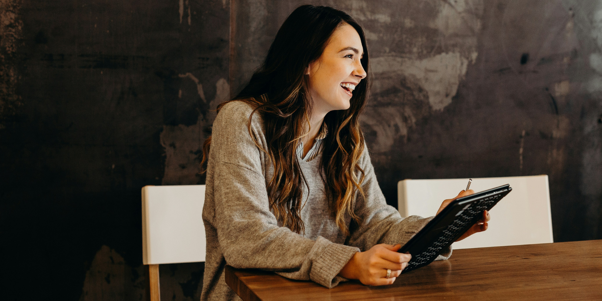 woman sitting at a table, holding a tablet and smiling enthusiastically